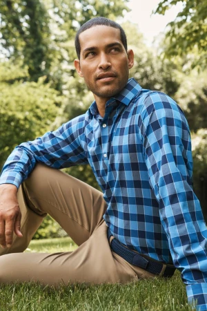 A man with short dark hair sits on green grass in a park, wearing a long-sleeved blue plaid shirt and khaki pants on a sunny day.