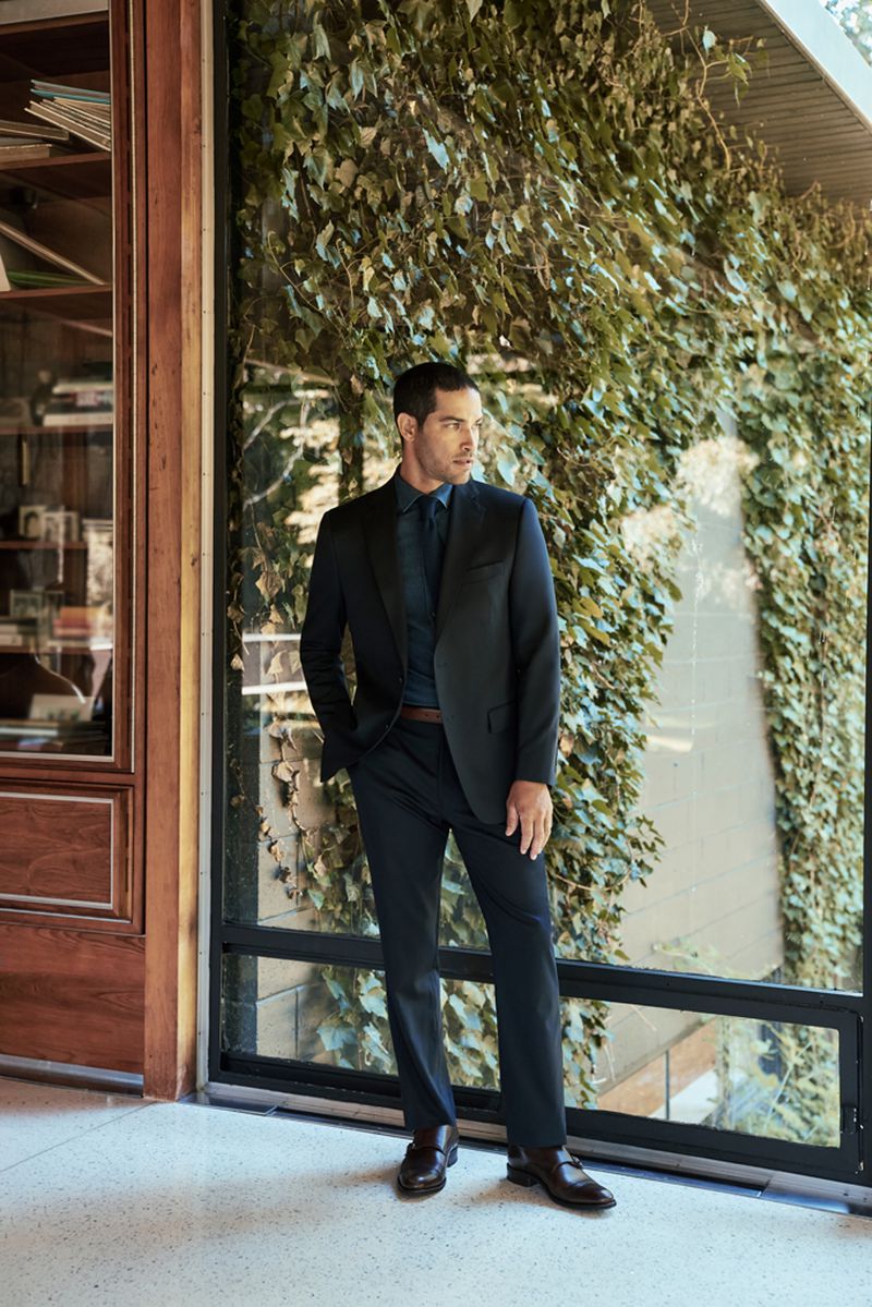 Man in a dark suit standing in front of a window covered with ivy. He is wearing a navy shirt, and dark brown shoes.