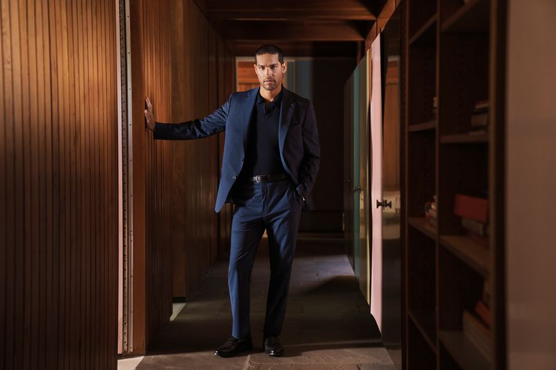 Portrait of a man in a blue suit standing in a wood-paneled hallway, hand resting on the wall, looking at the camera in a home interior.