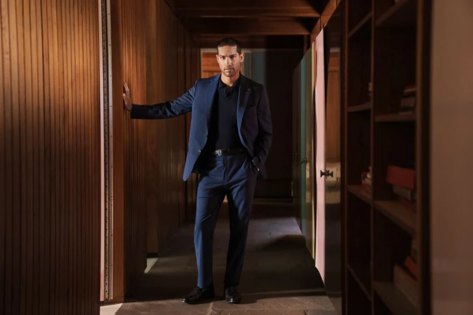 Portrait of a man in a blue suit standing in a wood-paneled hallway, hand resting on the wall, looking at the camera in a home interior.