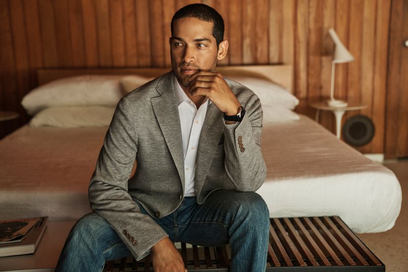 Adult male in blazer and jeans poses indoors sitting on a bench with bedroom background, looking off-camera with hand to chin.
