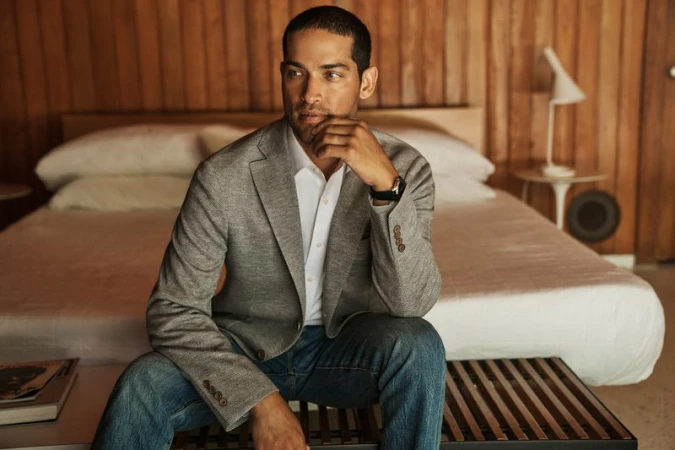 Man in a grey blazer and denim jeans seated on a wooden slat bench in front of a bed with wood-paneled walls.