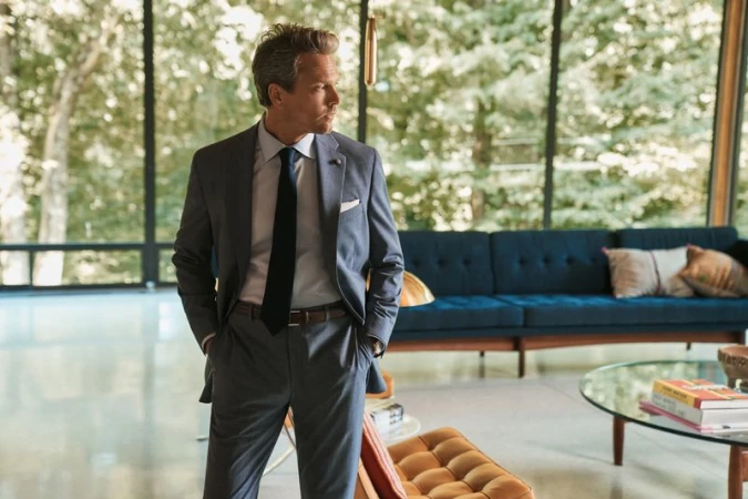 Adult man in a grey suit with a navy tie stands indoors with mid-century modern decor and floor-to-ceiling windows that reveal a leafy landscape.