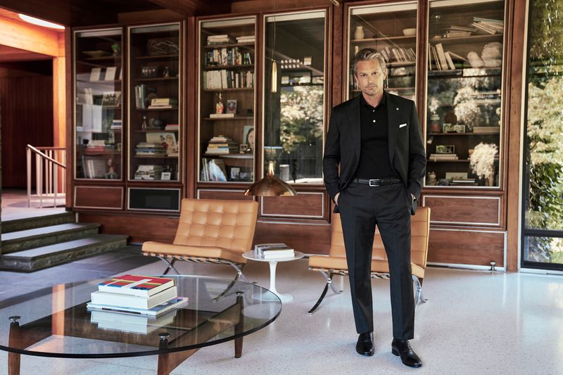 Man in black suit stands in mid-century modern interior with built-in bookshelves, tan leather chairs, and a glass coffee table.