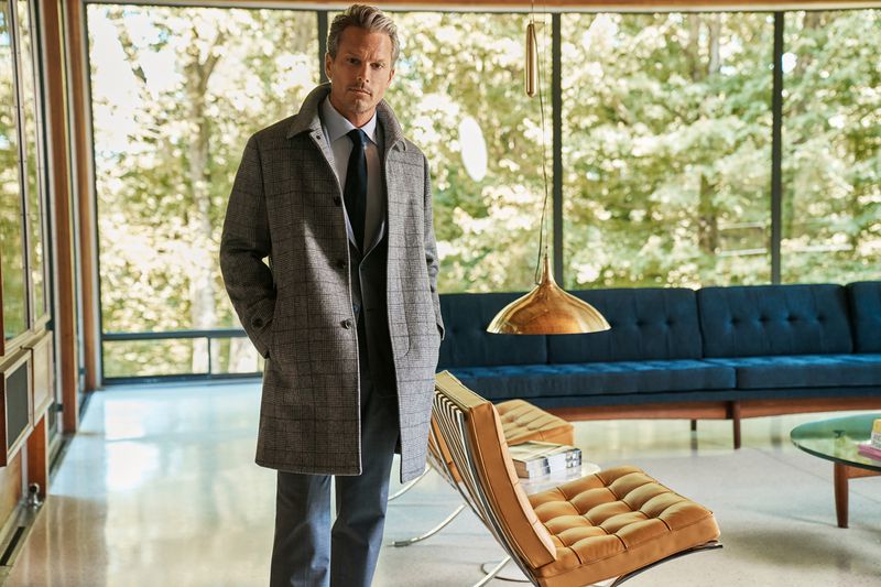 Adult man in a grey plaid coat, light blue dress shirt, and grey trousers poses in a modern home with natural outdoor lighting.