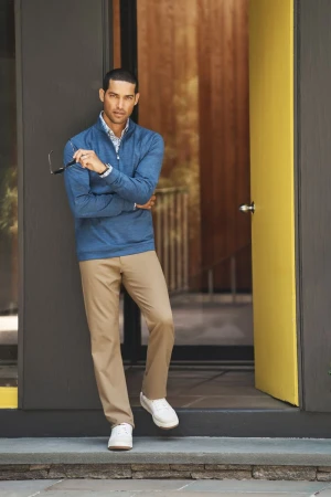 Adult male in blue quarter-zip shirt and khaki pants leaning against a modern doorway with sunglasses in hand looking at the camera.