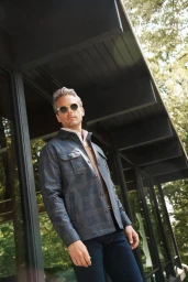 Man wearing sunglasses and a patterned blue shirt jacket standing outdoors against a modern architectural exterior.