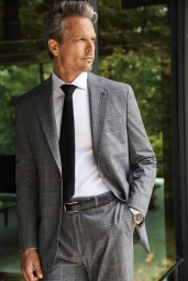 A man in a grey windowpane plaid suit, white shirt, and black tie poses near large glass architectural windows.