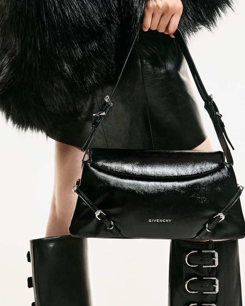 Model holding a glossy black Givenchy shoulder bag with silver buckle details, wearing black faux fur and boots against a white backdrop.
