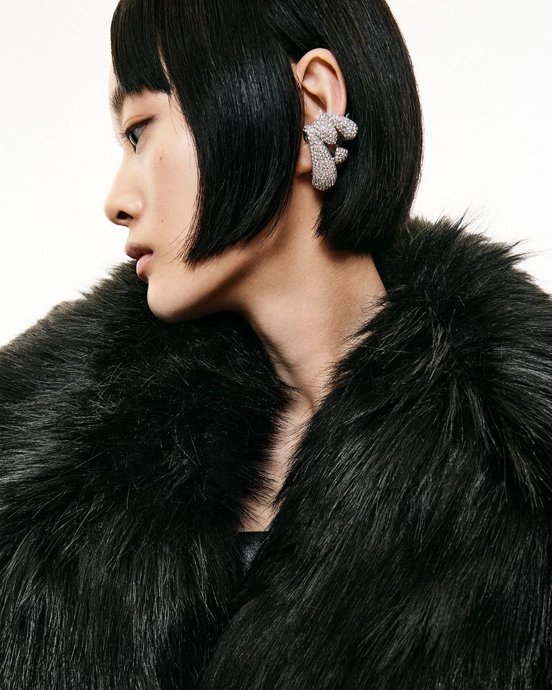 Profile shot of a young adult woman with black hair wearing a dark faux fur coat and a decorative silver ear cuff on a white background.