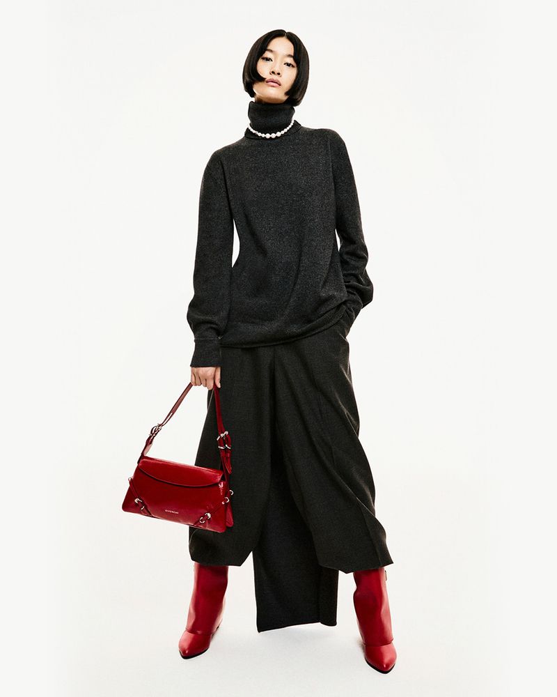 A full-length shot of an Asian female model posing in a dark gray sweater and culottes, red boots, and a red handbag against a white background.