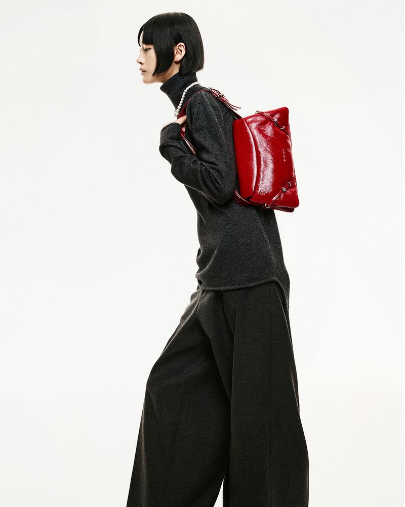 A slender person models a red leather bag in a fashion shot, wearing a dark grey turtleneck, pearl necklace, and wide-legged pants on white.