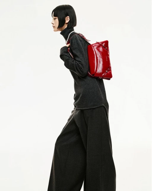 A slender person models a red leather bag in a fashion shot, wearing a dark grey turtleneck, pearl necklace, and wide-legged pants on white.