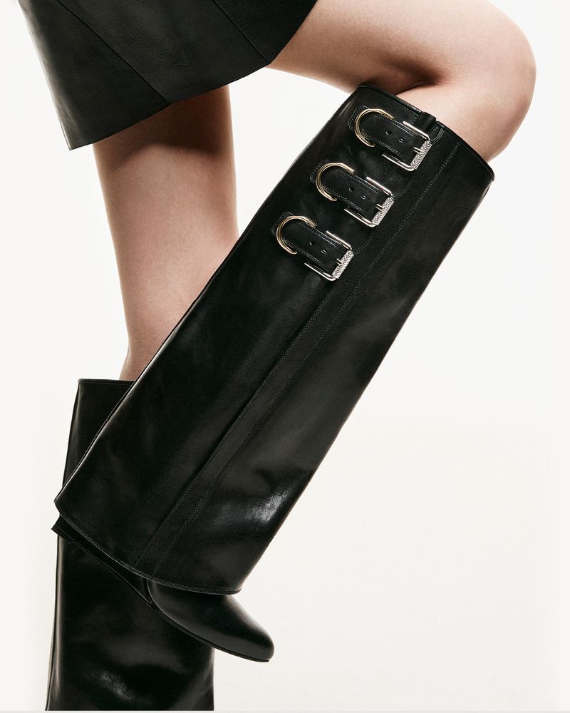 Close-up of a person wearing black leather knee-high boots with silver buckles, complemented by a matching black leather skirt.