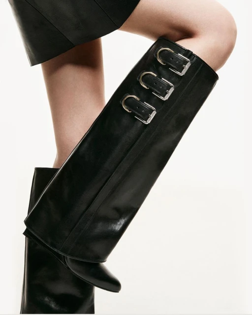 Close-up of a person wearing black leather knee-high boots with silver buckles, complemented by a matching black leather skirt.