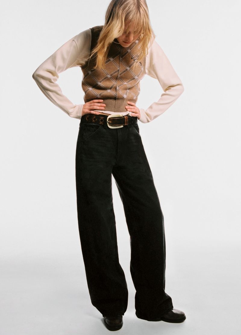 Blonde woman in a long-sleeved shirt, argyle sweater vest, dark wide leg jeans, and loafers posing in a photo studio with a white background.