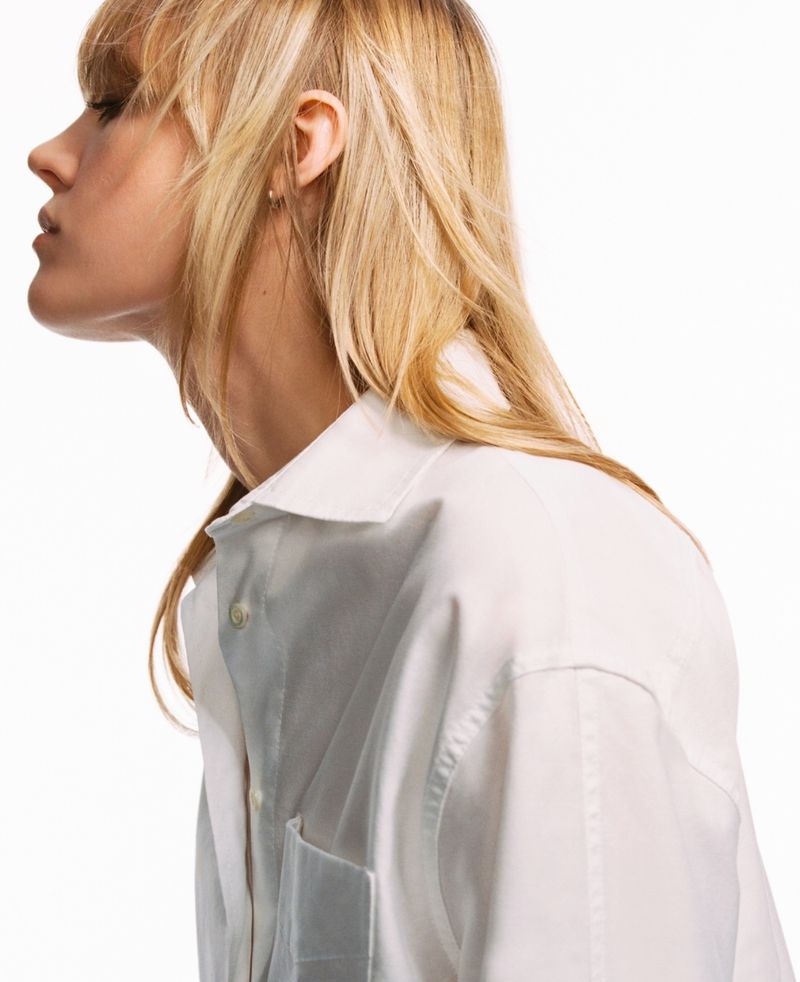 Profile of a blonde woman wearing a white button-down shirt against a white background, shot with studio lighting in portrait orientation.
