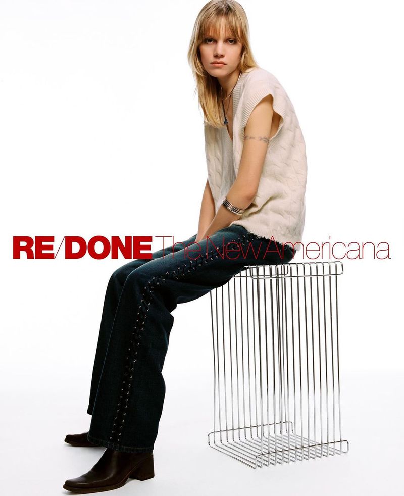 A blonde woman in a white sweater vest and dark jeans sits on a metal stool against a white backdrop in a fashion studio setting.