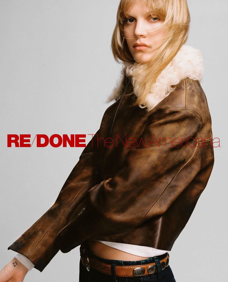 Blonde model in a brown leather jacket and jeans poses indoors against a neutral backdrop, showcasing a fashion editorial look.