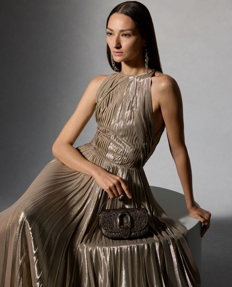 Elegant woman in a shimmering gold halter gown, seated and holding a beaded purse, illuminated by soft studio lighting.