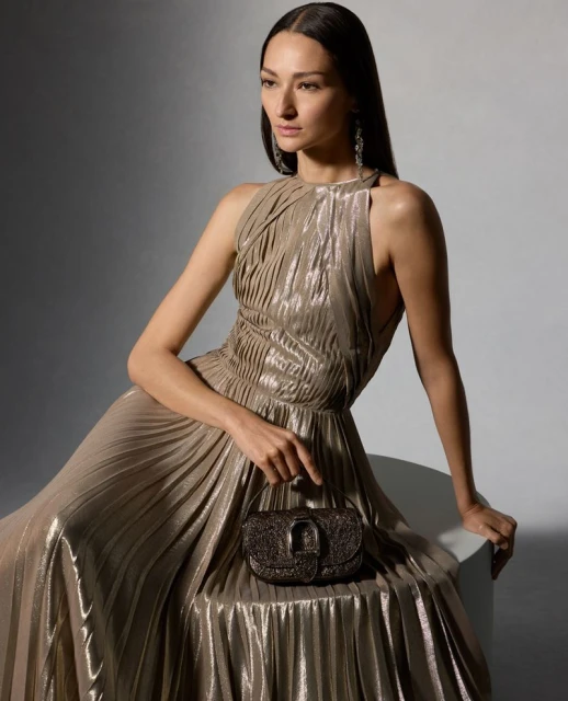 Elegant woman in a shimmering gold halter gown, seated and holding a beaded purse, illuminated by soft studio lighting.