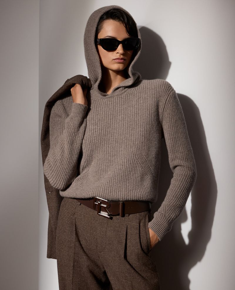 Adult woman wearing a hooded sweater, sunglasses, suit jacket, and pants stands indoors against a neutral wall in soft lighting.