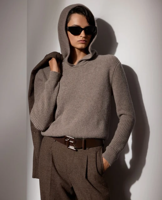 Adult woman wearing a hooded sweater, sunglasses, suit jacket, and pants stands indoors against a neutral wall in soft lighting.