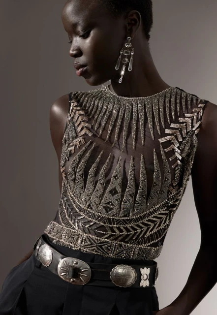 An African-American adult female fashion model poses in a sleeveless beaded top and black trousers with a silver concho belt.