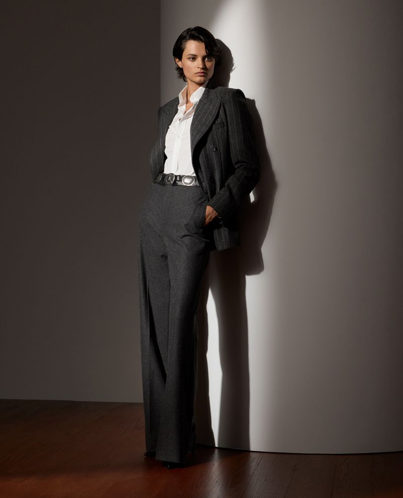 Fashion photo of a female model in a pinstripe suit leaning against a white wall. Indoor shot with studio lighting and shadow detail.