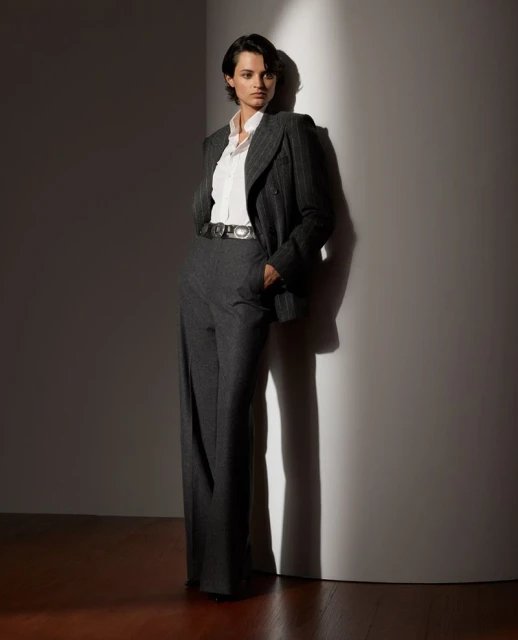 Fashion photo of a female model in a pinstripe suit leaning against a white wall. Indoor shot with studio lighting and shadow detail.