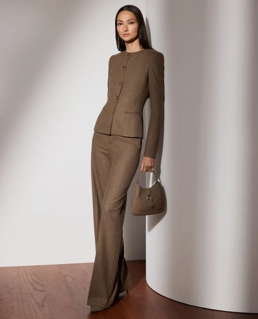 A woman in a brown suit leans against a curved wall with a brown handbag. The light creates shadows on the wall behind her.