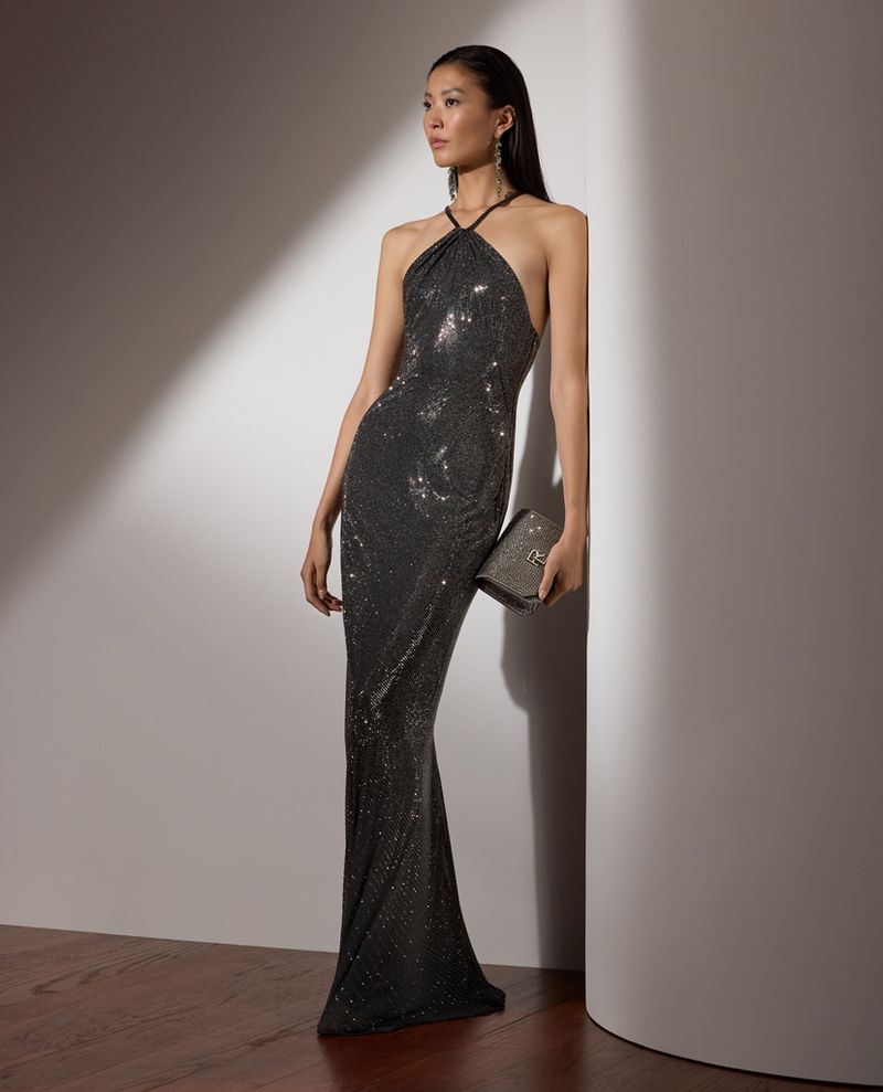 Elegant Asian woman in a sequined halter gown poses against a white wall, holding a clutch, with soft lighting and hardwood floors.