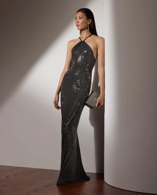 Elegant Asian woman in a sequined halter gown poses against a white wall, holding a clutch, with soft lighting and hardwood floors.