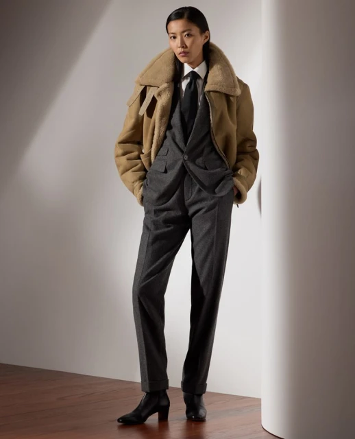 Asian woman in a grey suit, white shirt, and black tie poses indoors, wearing a beige shearling jacket and black heeled boots, standing in a stylish fashion pose.