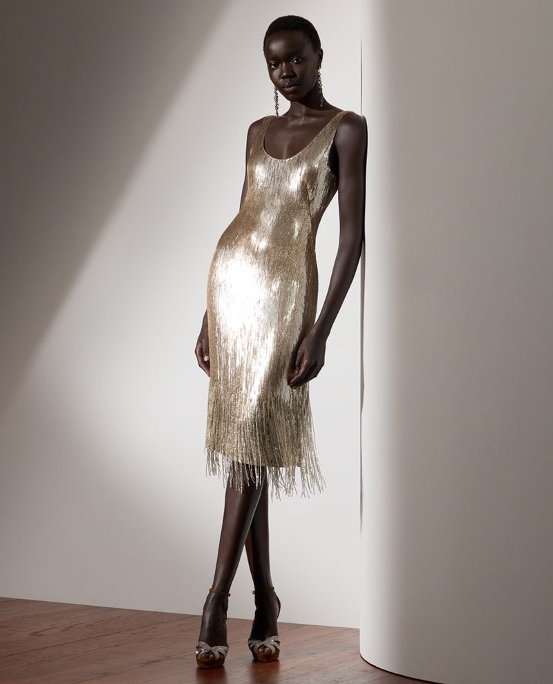 Elegant dark-skinned woman poses in a gold fringed dress and strappy heels, leaning against a neutral wall with soft lighting.