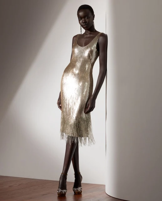 Elegant dark-skinned woman poses in a gold fringed dress and strappy heels, leaning against a neutral wall with soft lighting.