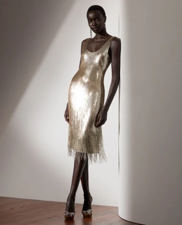 Elegant dark-skinned woman poses in a gold fringed dress and strappy heels, leaning against a neutral wall with soft lighting.