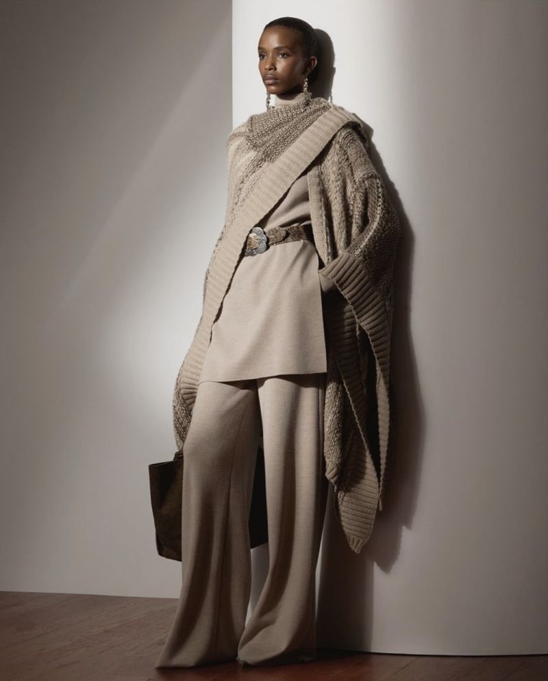 Elegant Black female fashion model showcases beige knitwear shawl, top, and wide-leg pants against a neutral background in a studio setting.