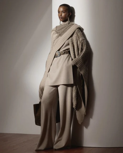 Elegant Black female fashion model showcases beige knitwear shawl, top, and wide-leg pants against a neutral background in a studio setting.