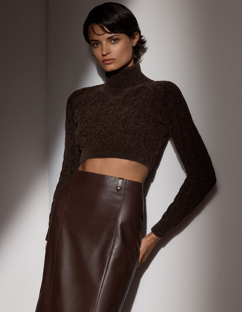 Portrait of a brunette woman wearing a brown knit top and leather midi skirt, posing in front of a neutral grey background with studio lighting.