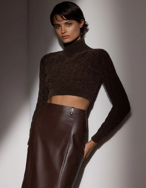 Portrait of a brunette woman wearing a brown knit top and leather midi skirt, posing in front of a neutral grey background with studio lighting.