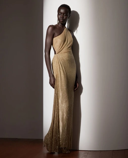 Elegant Black woman poses in a shimmering gold, one-shoulder gown, showcasing a blend of classic style and modern fashion in a studio setting.