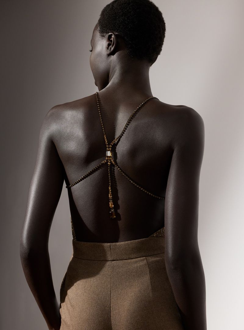 Dark-skinned woman with bare back in gold beaded top and brown pants in a medium shot, studio setting with soft lighting.