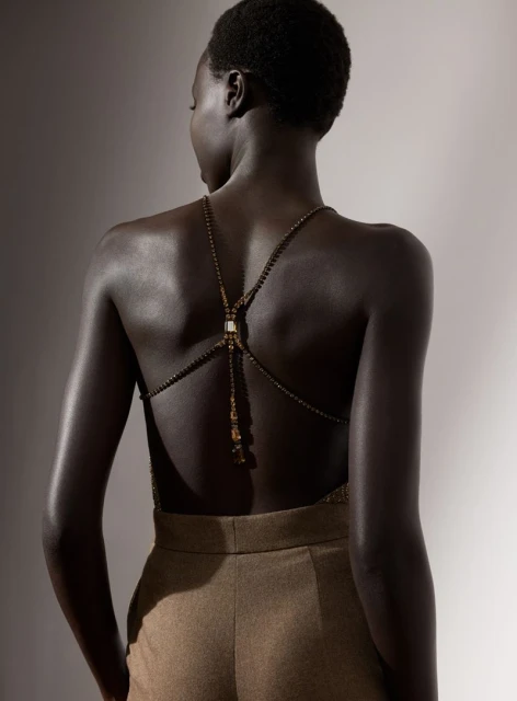 Dark-skinned woman with bare back in gold beaded top and brown pants in a medium shot, studio setting with soft lighting.