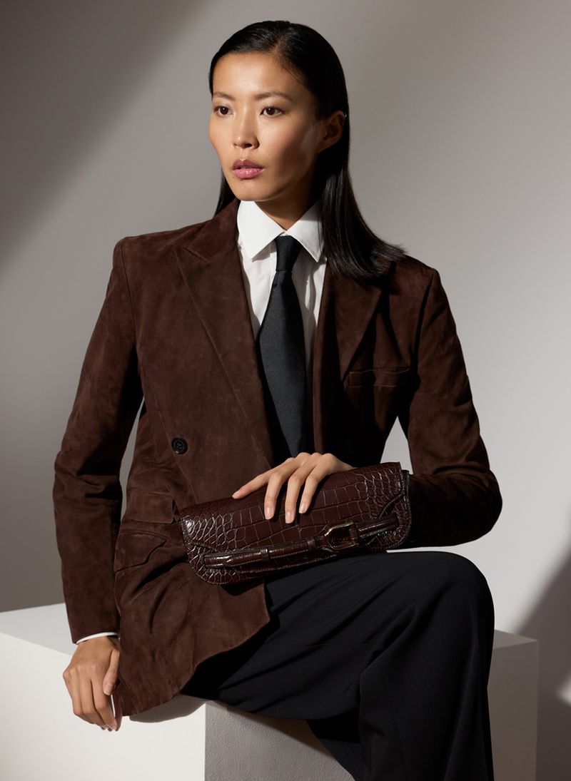 Elegant Asian woman in brown jacket and black tie sits, holding crocodile clutch against a light background with soft shadows.