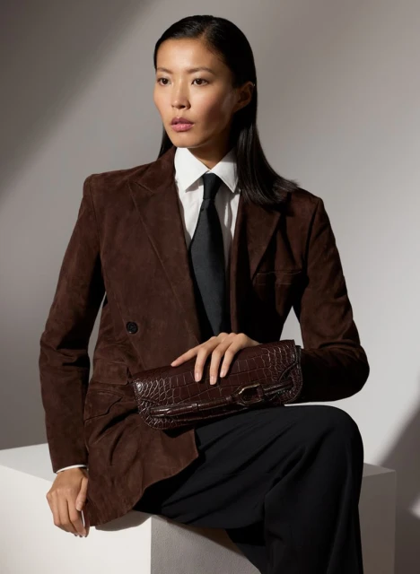 Elegant Asian woman in brown jacket and black tie sits, holding crocodile clutch against a light background with soft shadows.