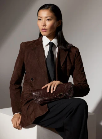 Elegant Asian woman in brown jacket and black tie sits, holding crocodile clutch against a light background with soft shadows.