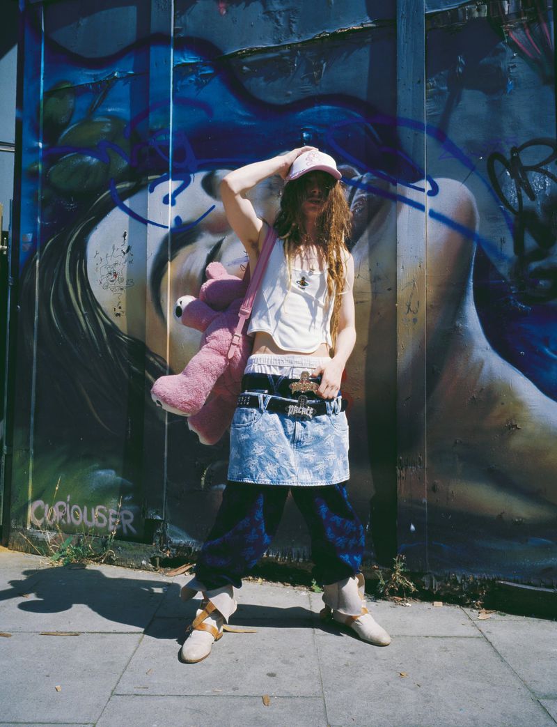 Adult wearing crop top, cap, and denim mini skirt poses against an urban wall covered in graffiti art with a pink teddy bear.