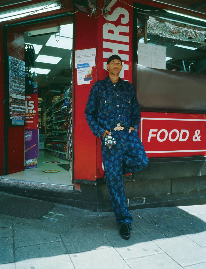 Man in a blue patterned suit standing outside a convenience store with red and white signage on a sunny day in an urban setting.