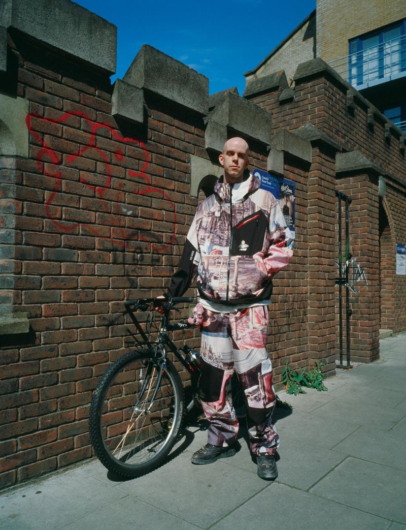 A bald man in a patterned jacket and pants stands with a bicycle in front of a brick wall on a city sidewalk on a sunny day.